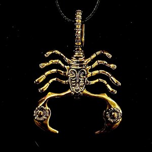 K&ouml;nnte beinhalten: Eine goldfarbene Skorpion-Anh&auml;nger-Halskette. Der Skorpion ist mit komplizierten Schnitzereien verziert und hat eine schwarze Schnur.