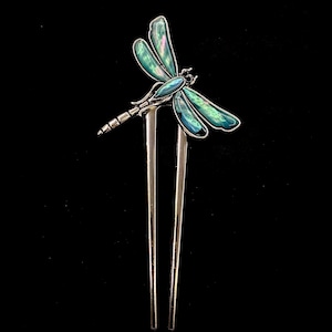 May include: A silver-toned hair pin featuring a dragonfly design. The dragonfly has iridescent teal and blue wings and a detailed body. The pin has two long, parallel prongs, perfect for securing hair. The background is black.