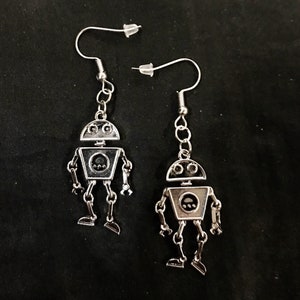 May include: A pair of silver-colored robot earrings. Each earring features a robot design with a rounded head, square body, and articulated limbs. The earrings hang from silver-colored fish hook ear wires with clear rubber stoppers.