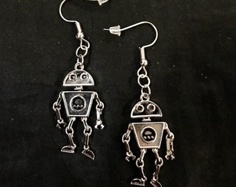 May include: A pair of silver robot earrings. Each earring has a robot design with a rounded head, square body, and articulated limbs. The earrings hang from silver fish hook ear wires with clear rubber stoppers.