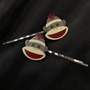 May include: Two silver hair clips with monkey designs. Each monkey is wearing a red and white knit hat. The monkeys have brown fur and white faces.