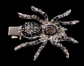 May include: A silver-toned spider hair clip featuring a metallic body encrusted with small black and clear crystals. The spider has eight articulated legs and a smooth, dark oval-shaped stone set in the center of its back, attached to a metal clip.