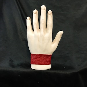 May include: A white ceramic hand with a red patterned bracelet. The hand is raised with fingers spread apart.