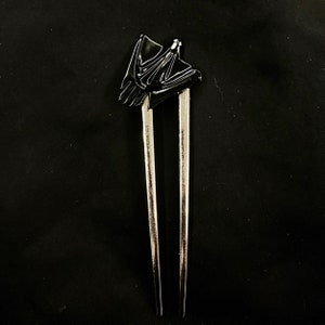 May include: A silver hair stick with a black decorative top. The hair accessory has two long, parallel prongs. The top is a stylized, abstract shape with a glossy finish, set against a dark background.