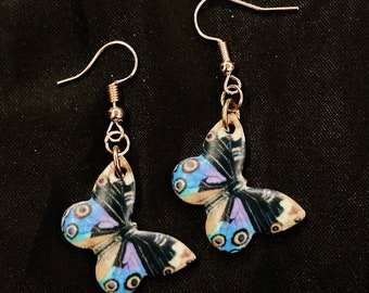 May include: A pair of butterfly-shaped earrings. Each earring features a detailed butterfly design with blue, purple, and tan wings. The earrings have gold-colored fish hook ear wires and are set against a black background.