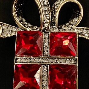 May include: A silver-toned brooch in the shape of a gift box with a red bow. The box is decorated with clear rhinestones and has four red, square-shaped gems.