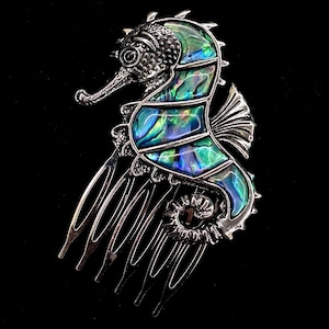 May include: A silver-toned seahorse hair comb with iridescent abalone shell inlay. The seahorse design features detailed textures and a comb with multiple tines. The background is black.