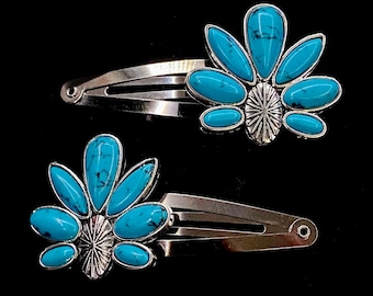 May include: Two silver-toned hair barrettes, each adorned with a turquoise-colored stone arrangement. The stones are oval and teardrop-shaped, set around a central, sunburst-like design. The background is solid black.