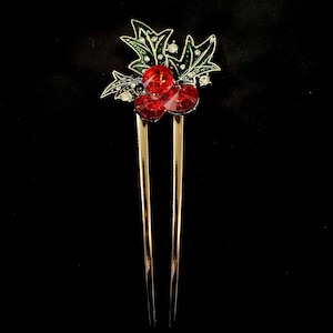 May include: A gold-toned hair pin featuring a holly design. The pin has three large red jewel-like berries and green and white enamel leaves. The pin is set against a black background.