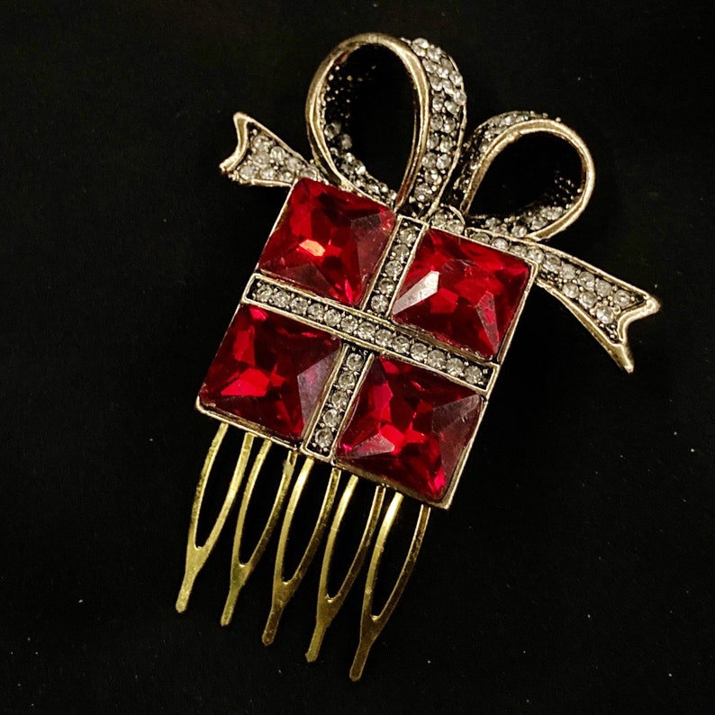 May include: A gold-tone hair comb with a red rhinestone gift box design. The comb has a bow and a series of teeth on the bottom.