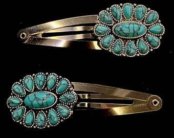 May include: Two silver-toned hair clips, each adorned with a turquoise-colored stone design. The design features a central oval stone surrounded by smaller, teardrop-shaped stones, creating a floral pattern. The clips are set against a black background.
