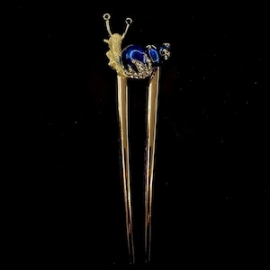 May include: Gold hair stick with a blue and gold snail design. The snail has rhinestones on its shell.