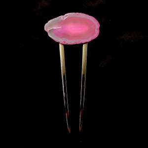 May include: A pink and gray agate hair pin with two gold-colored prongs. The agate stone is oval-shaped and has a polished surface. The hair accessory is set against a black background.