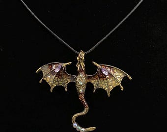 May include: A gold dragon pendant necklace with purple and iridescent gemstones. The dragon is in flight with its wings spread wide. The necklace is on a black cord.