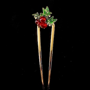 May include: A gold-toned hair pin with a decorative top featuring red jewel-like berries and green leaves with white accents. The hair accessory is set against a black background.