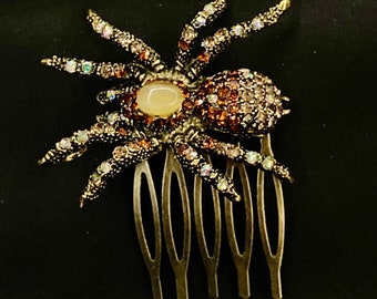 May include: A gold-tone spider hair comb with iridescent rhinestones and a large, off-white faux pearl in the center. The comb has seven teeth.