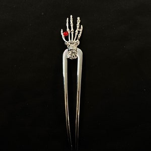 May include: Silver hair fork with a skeleton hand design and a red rhinestone on the index finger.