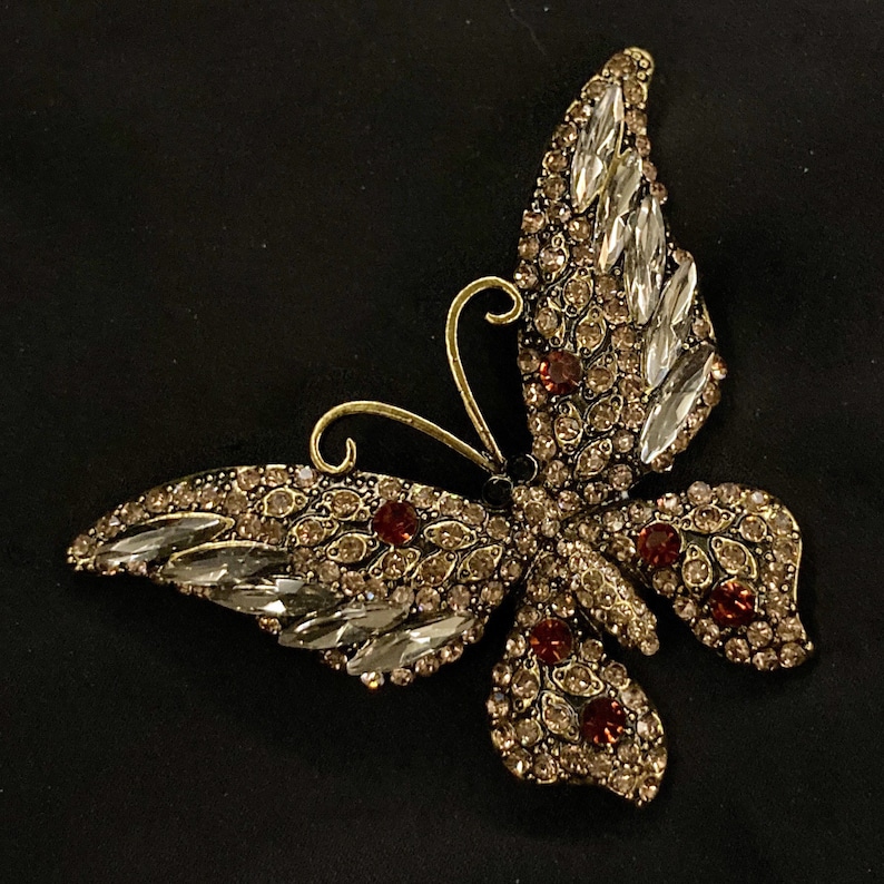Puede incluir: Un broche de mariposa de tono dorado con diamantes de imitaci&oacute;n transparentes y marrones. La mariposa tiene un dise&ntilde;o detallado y es una pieza llamativa.