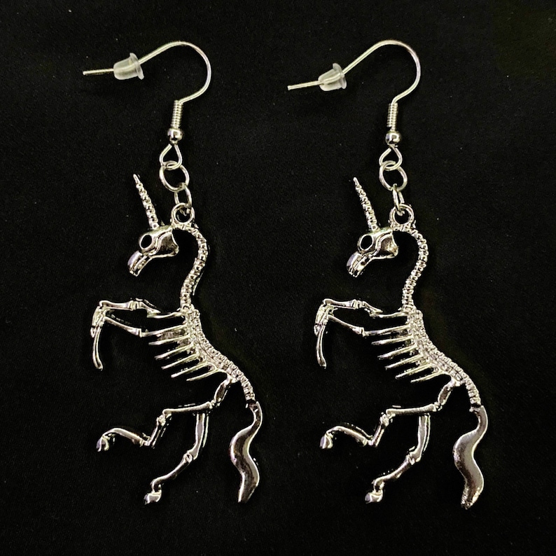 May include: A pair of silver skeleton unicorn earrings. The earrings are dangling and have a hook closure.