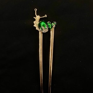 May include: A gold hair stick with a green snail design. The snail has a green shell and gold accents.