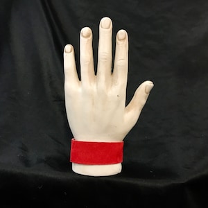 May include: A white ceramic hand with five fingers raised, wearing a red velvet wrist cuff.
