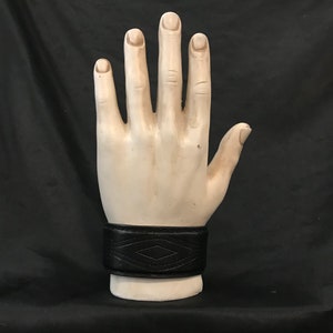 May include: A black leather wristband with a diamond-shaped pattern. The wristband is worn on a plaster hand model.