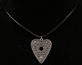 Könnte beinhalten: Silberfarbene herzförmige Anhänger-Halskette mit Ouija-Brett-Design. Der Anhänger hat eingravierte Buchstaben, Zahlen und dekorative Elemente, mit einem kleinen Totenkopf am unteren Ende. Die Halskette hängt an einem schwarzen Band.