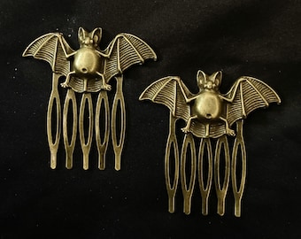 Op de afbeelding: Twee gouden metalen haar kammen in de vorm van een vleermuis. Elke kam heeft een gedetailleerd vleermuisontwerp met een textuur. De kammen hebben elk zeven tanden.