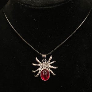 Puede incluir: Un collar con colgante de araña en tono plateado que presenta una piedra preciosa roja oscura de forma ovalada engastada en el abdomen. La araña tiene ocho patas texturizadas y cuelga de un cordón negro fino y sencillo alrededor del cuello.