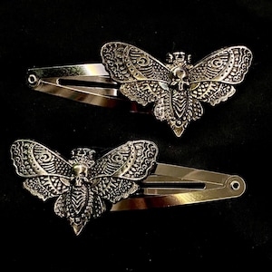 May include: Two silver hair clips featuring a moth design. The moths have detailed wing patterns and a skull motif. The clips are set against a black backdrop.