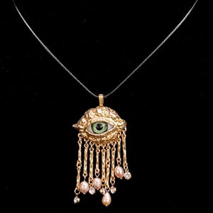 May include: Gold-toned pendant necklace featuring an eye design with a green iris and white sclera. The pendant has a textured surface and is adorned with dangling chains, pink pearls, and small clear crystals. The necklace hangs on a thin, dark cord.