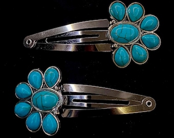 May include: Two silver-toned hair clips with turquoise-colored stone flower designs. Each clip features an oval center stone surrounded by teardrop-shaped stones, set against a black background. These hair accessories are suitable for various hairstyles.