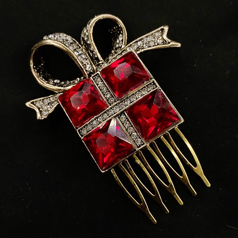 May include: A gold-tone hair comb featuring a red jeweled gift box design. The gift box has a bow and is decorated with clear rhinestones.