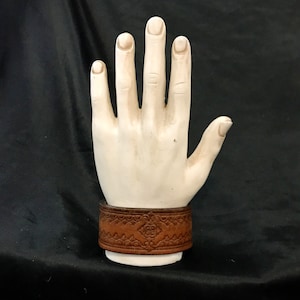 May include: A brown leather bracelet with an intricate embossed design. The bracelet is on a white hand model.