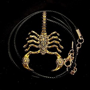 K&ouml;nnte beinhalten: Eine goldfarbene Skorpion-Anh&auml;ngerkette mit einem schwarzen Lederband. Der Skorpion ist mit aufwendigen Schnitzereien verziert und hat ein florales Design am Schwanz.
