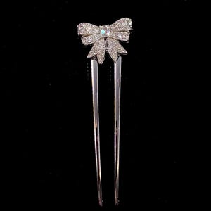 May include: A silver-toned hair fork featuring a decorative bow at the top. The bow is encrusted with numerous small, clear, sparkling rhinestones and has a single, larger, rectangular-cut clear stone centered in the middle of the bow design.