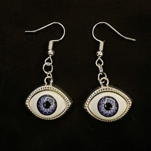 May include: A pair of silver eye earrings with a white background and a purple iris. The earrings have a silver hook and a chain.