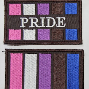 Op de afbeelding: Twee bruine stoffen patches met geborduurde strepen in roze, wit, paars, bruin en blauw. De bovenste patch heeft het woord "PRIDE" in wit geborduurd.