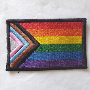 Peut inclure: Un patch de drapeau arc-en-ciel avec une bordure noire et un triangle brun, bleu, bleu clair, rose et blanc dans le coin.