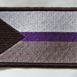 May include: An embroidered patch featuring a brown triangle, a white stripe, a purple stripe and a brown stripe. The patch has a brown border.
