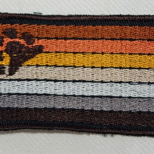 May include: An embroidered patch featuring a brown bear paw print in the centre and seven horizontal stripes in shades of brown, orange, yellow, beige, grey, and white. The patch has a black border.