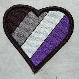 May include: An embroidered heart-shaped patch featuring the asexual pride flag colours: black, grey, white, and purple.