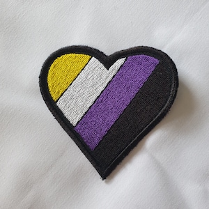 May include: A heart-shaped patch with the colors of the non-binary pride flag. The patch is yellow, white, purple, and black.