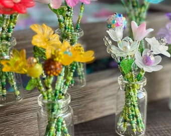 May include: A collection of miniature glass bottles containing small bouquets of artificial flowers crafted from colorful beads and translucent plastic petals. Each stem is made of green seed beads, and the flowers feature various colors including red, yellow, pink, blue, orange, and purple.