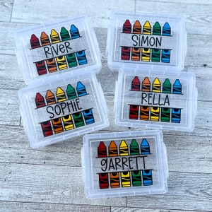 May include: Five clear plastic boxes with lids, each labeled with a name and a crayon design. The crayon design features a row of six crayons in red, orange, yellow, green, and blue. The names are River, Simon, Sophie, Rella, and Garrett.