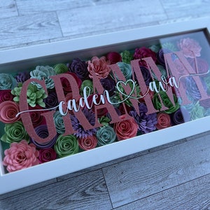 May include: A framed floral art piece with a white border. The artwork features paper flowers in shades of pink, purple, and green. The names "GRAM" and "ava" are displayed in a glittery pink font, with "caden" in a cursive font.