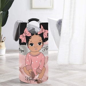 May include: A cylindrical laundry hamper featuring a cartoon illustration of a baby girl with pink bows and a pink polka-dot outfit. The hamper has a black handle and the name "Harleyn" is printed on it. The background is a light pink tufted design.