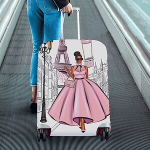 May include: A rolling suitcase with a pink and black illustration of a woman in a pink dress in front of the Eiffel Tower. The suitcase has a white background and black wheels. The woman is wearing sunglasses and carrying a pink purse.