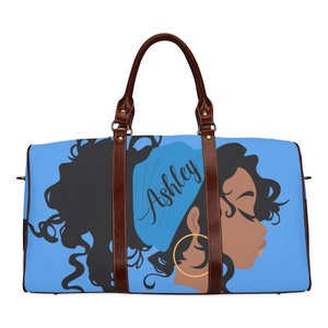 May include: A blue duffel bag featuring a graphic of a woman's profile with black hair, a blue headscarf, and gold hoop earrings. The name "Ashley" is written on the headscarf in cursive. The bag has brown leather handles and accents.