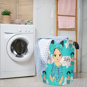 May include: A turquoise laundry hamper featuring a cartoon baby design with a bow and the name "Lordes". The hamper is filled with a blue and white star-patterned blanket. A washing machine and towels are in the background.